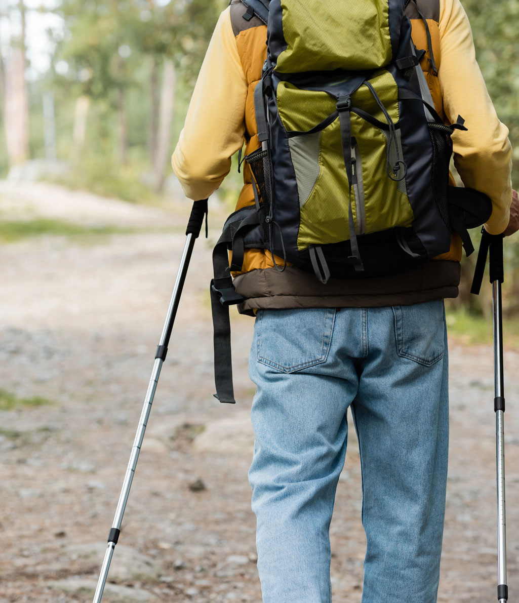 Vista posteriore di un escursionista con pantaloncini color cachi e una maglia scura, che sale un ripido sentiero di montagna sterrato con un grande zaino blu e nero e un bastone da trekking. Alberi sparsi e montagne lontane sono visibili sotto un cielo chiaro."