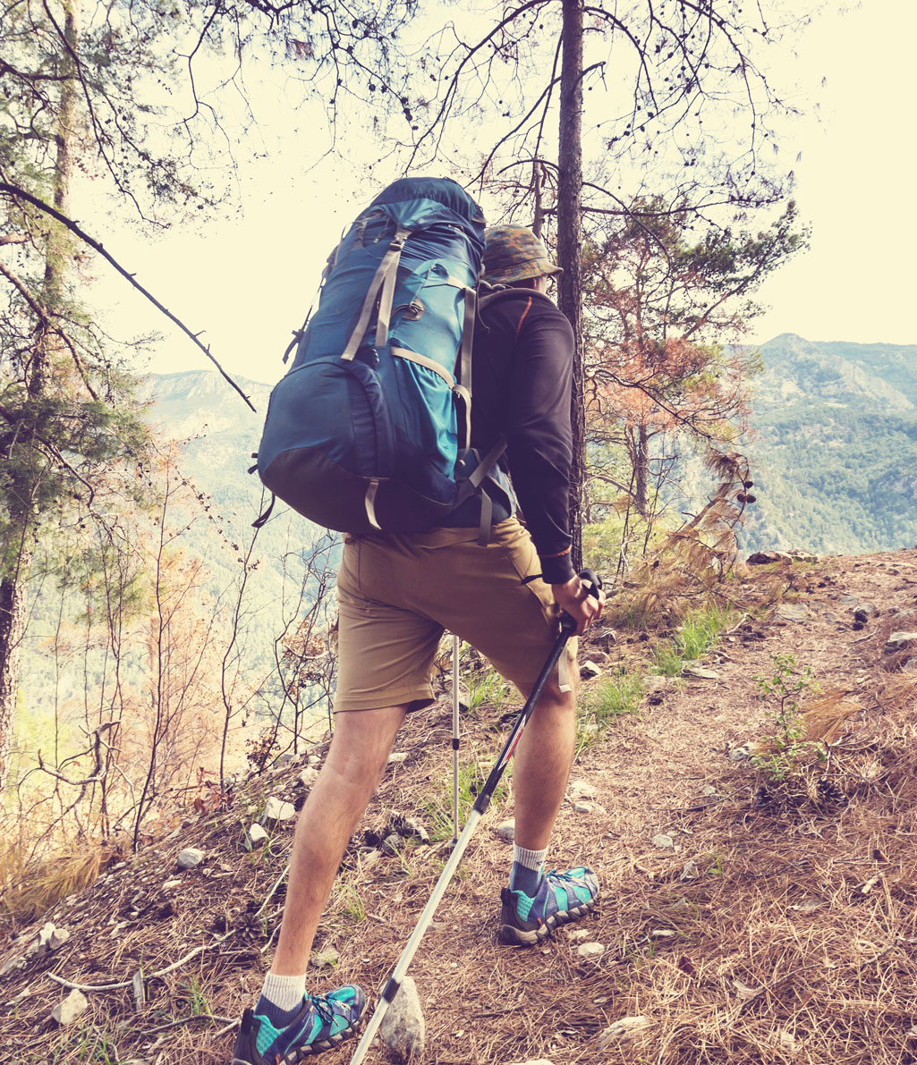 Vista posteriore di una persona che indossa una giacca gialla e jeans blu chiaro, con un grande zaino verde e nero da trekking e che usa due bastoni da passeggio su un sentiero sterrato in una foresta.