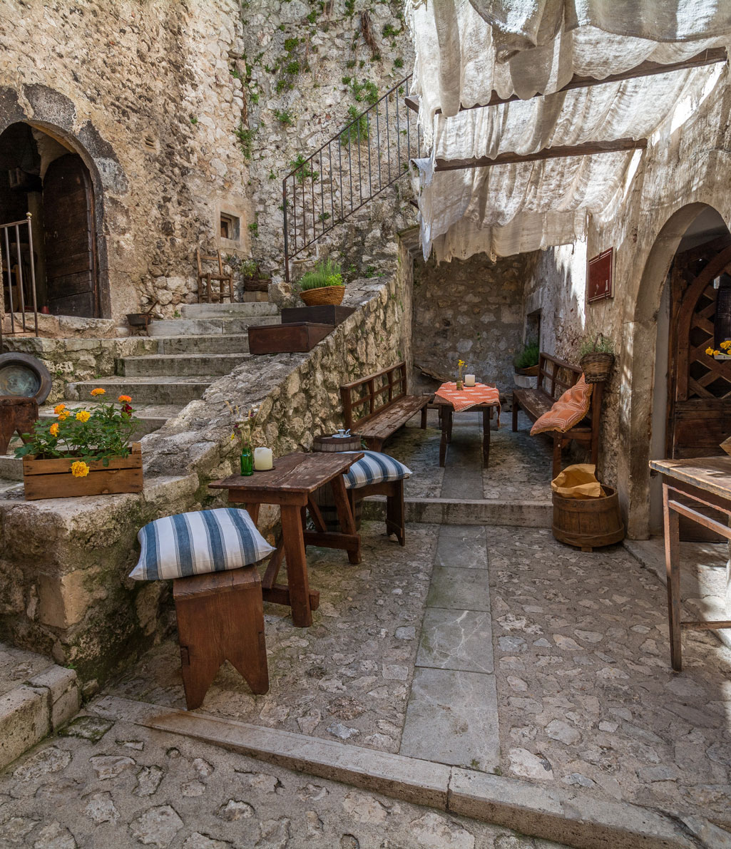 Un pittoresco cortile in pietra con tavoli e panche di legno, scale in pietra che conducono a un portone ad arco, piante in vaso con fiori arancioni e un telo bianco sospeso che fornisce ombra. L'ambiente è rustico e tradizionale.