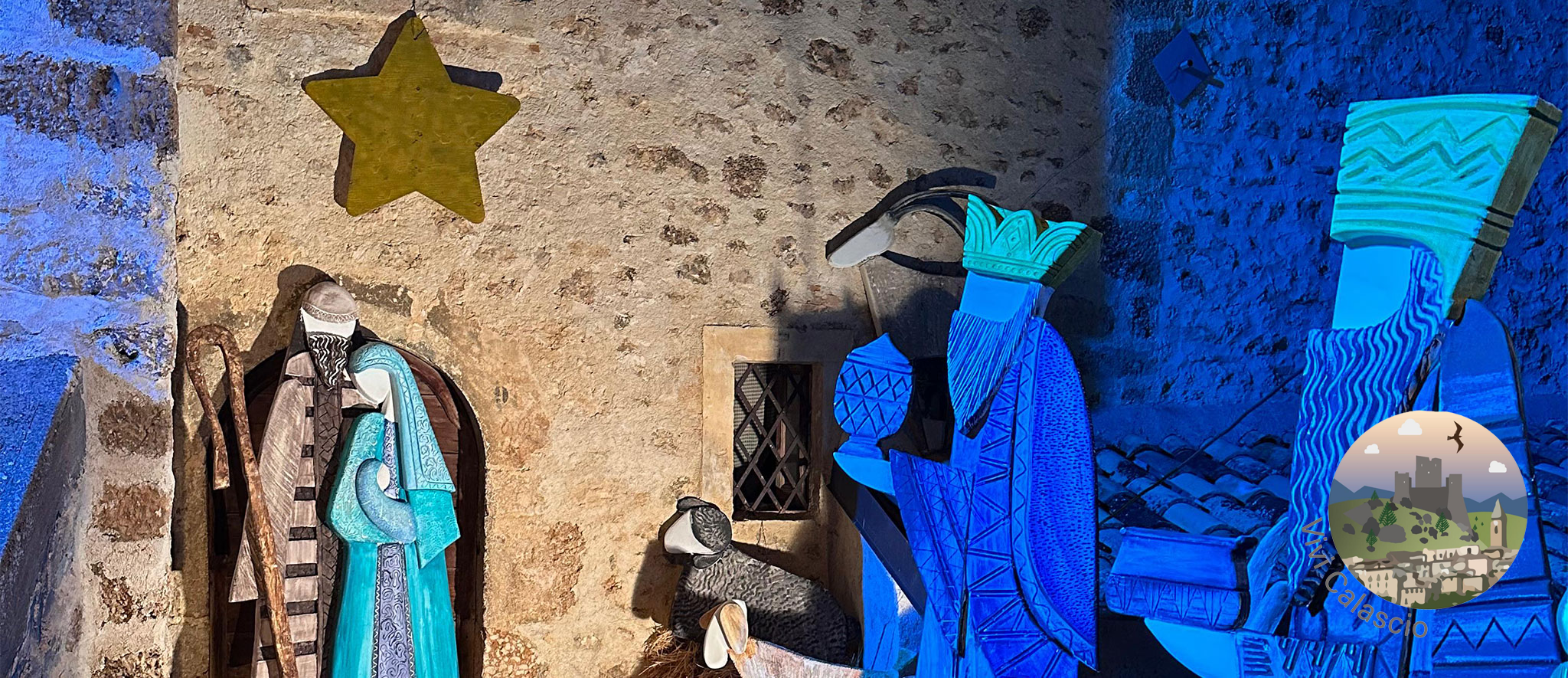 Una scena della natività all'aperto, con grandi figure stilizzate ritagliate in legno dipinto. A sinistra, la Madonna e San Giuseppe (o figure simili) sono rappresentate in toni di azzurro e grigio e sono illuminate da una luce calda giallo-oro. Al centro, un animale stilizzato in grigio scuro. A destra, i Re Magi sono rappresentati in un vibrante blu cobalto e beige, illuminati da una forte luce blu che crea un contrasto drammatico. Un'enorme stella gialla a cinque punte è appesa in alto a sinistra, brillando sopra la scena. Le figure sono ambientate contro una parete di pietra antica e irregolare con una piccola finestra rettangolare. Nell'angolo in basso a destra è visibile un logo circolare con la scritta 'Vivi Calascio' e un'illustrazione di un castello e uccelli.