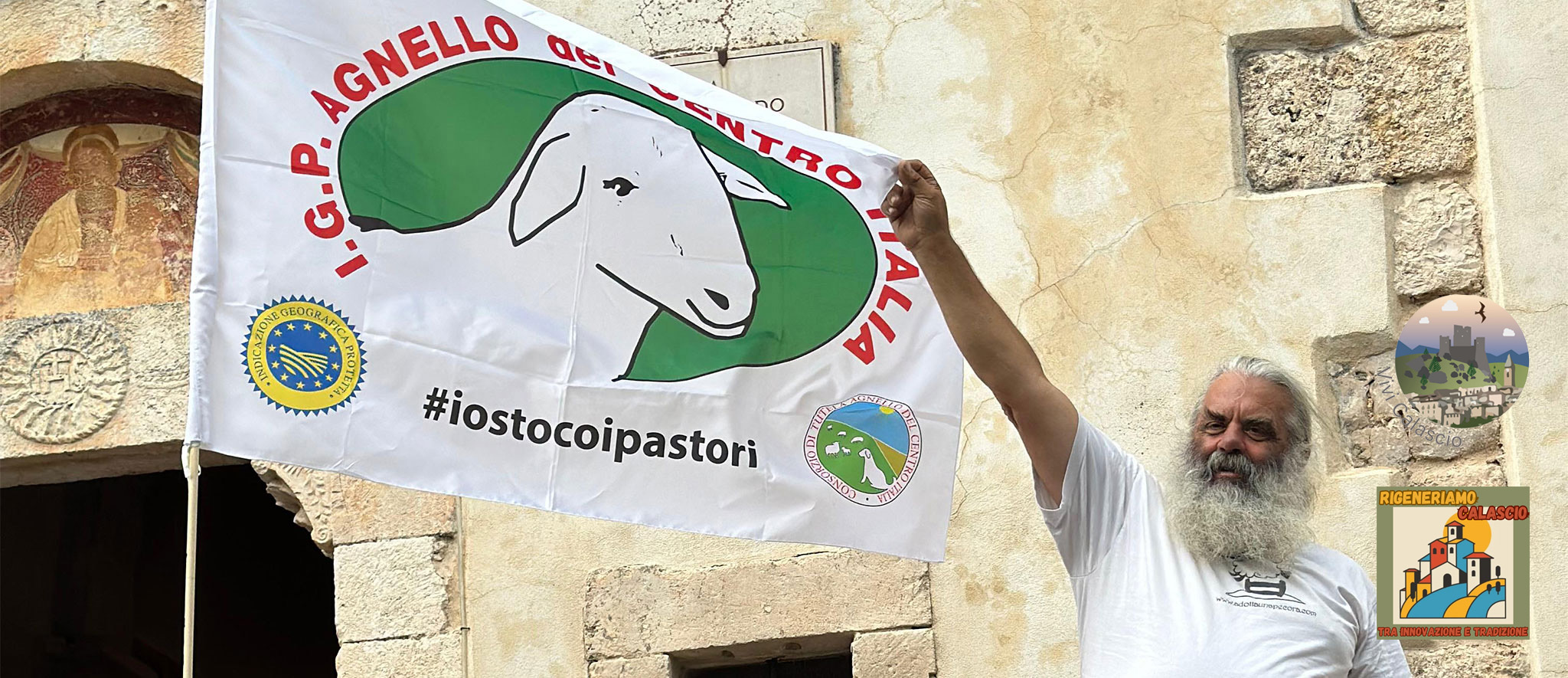 Un uomo anziano con una lunga barba bianca e capelli bianchi, che indossa una maglietta bianca, tiene alta una grande bandiera bianca. La bandiera mostra una sagoma verde di una testa di agnello e le scritte in rosso e nero "I.G.P. AGNELLO DEL CENTRO ITALIA" e l'hashtag nero "#iostocoipastori". Sono visibili anche il logo europeo I.G.P. giallo e blu e un altro logo circolare con pecore e montagne. L'uomo si trova davanti a un antico edificio in pietra con un ingresso ad arco e affreschi sbiaditi sulla sinistra. Nell'angolo in alto a destra, sono presenti altri loghi che promuovono iniziative locali, tra cui "RIGENERIAMO CALASCIO.