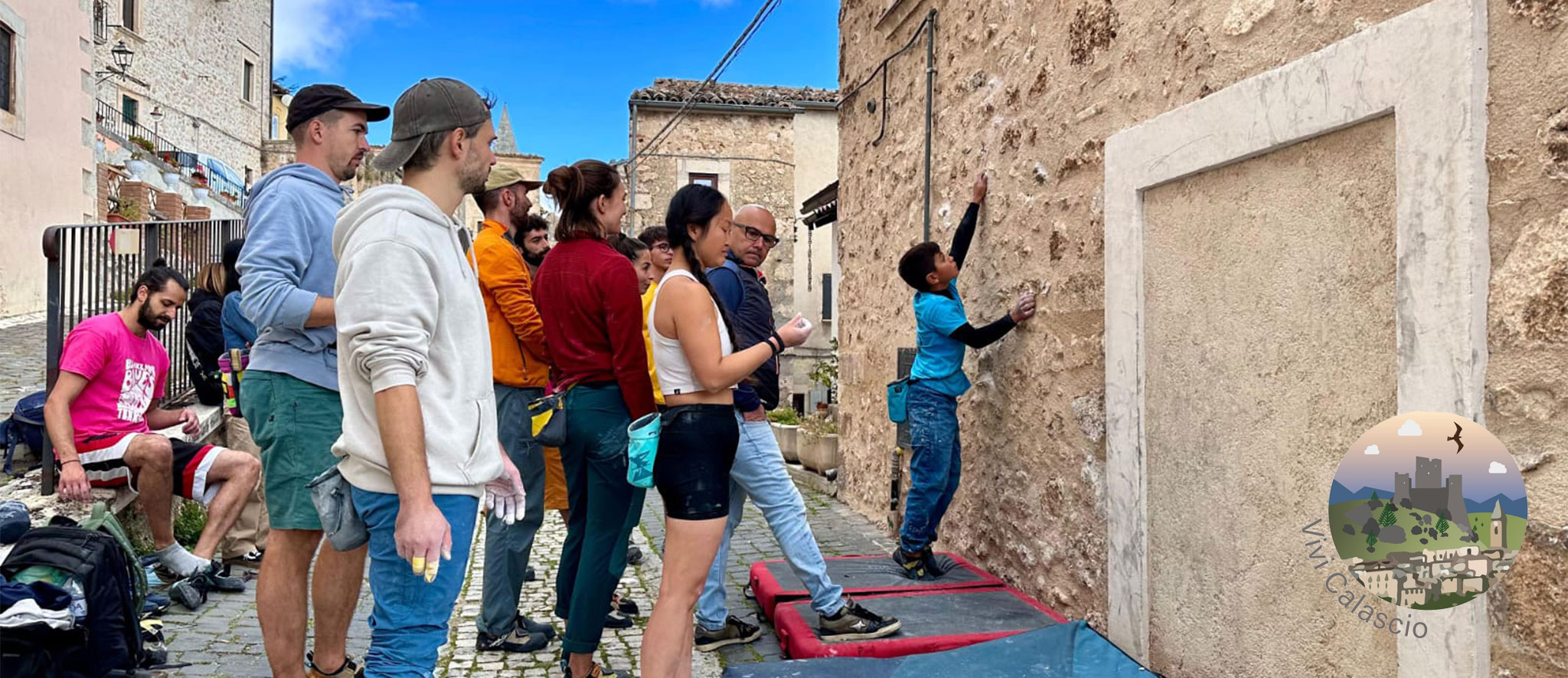 Un giovane bambino in una maglietta blu brillante sta facendo bouldering su una bassa parete di arrampicata in pietra chiara, incorporata in un edificio antico. Un gruppo di circa otto adulti, vestiti con abiti casual da esterno (tra cui felpe, magliette colorate e pantaloni sportivi), osserva il bambino da vicino sul lato sinistro dell'immagine. Sono presenti dei crash pad blu alla base della parete per ammortizzare le cadute. L'ambientazione è una stretta via acciottolata di un villaggio con edifici storici in pietra sullo sfondo. Nell'angolo in basso a destra è visibile un logo circolare con la scritta 'Vivi Calascio' e un'illustrazione di un castello e uccelli.