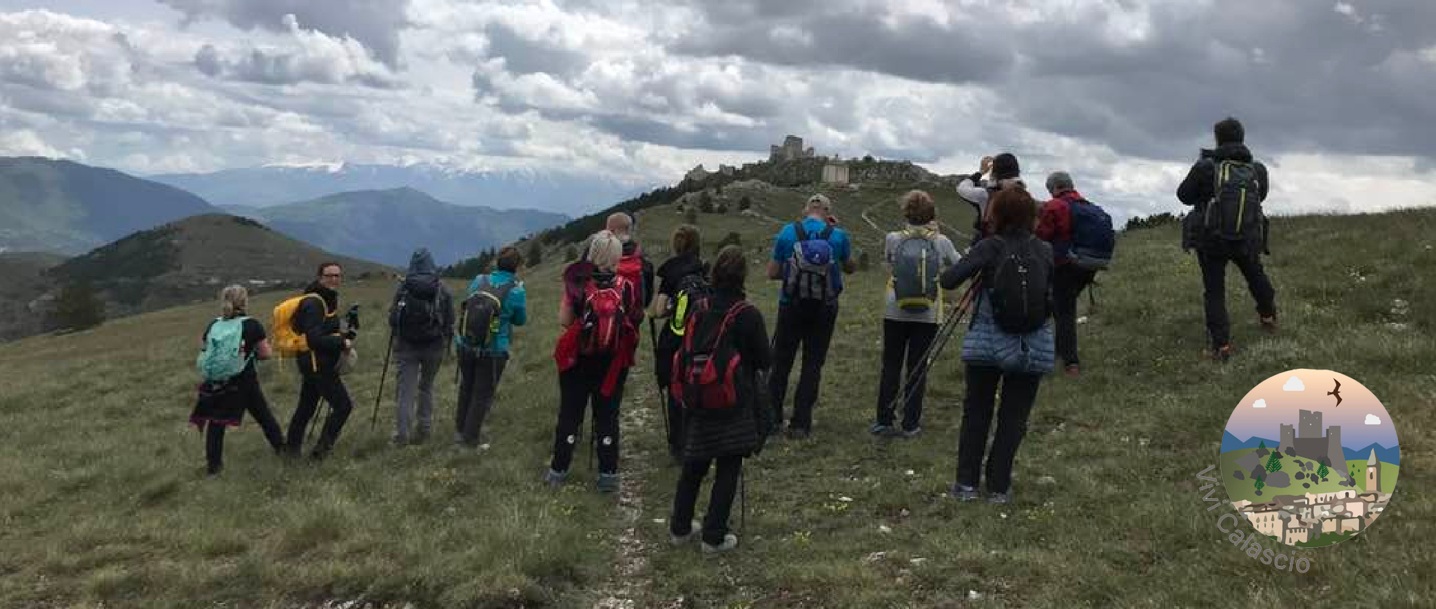 Un gruppo di circa dieci escursionisti, molti con zaini colorati (rossi, blu, gialli) e bastoni da trekking, sono in piedi su un pendio erboso, la maggior parte visti da dietro, osservando le imponenti rovine di un antico castello di pietra (il Castello di Rocca Calascio) in cima a una collina in lontananza. Il paesaggio circostante è caratterizzato da valli verdi e montagne che si estendono all'orizzonte, alcune con cime ancora innevate sotto un cielo nuvoloso e drammatico. Il terreno in primo piano è coperto da erba verde e secca. Nell'angolo in basso a destra è visibile un logo circolare con la scritta 'Vivi Calascio' e un'illustrazione di un castello e uccelli in volo.