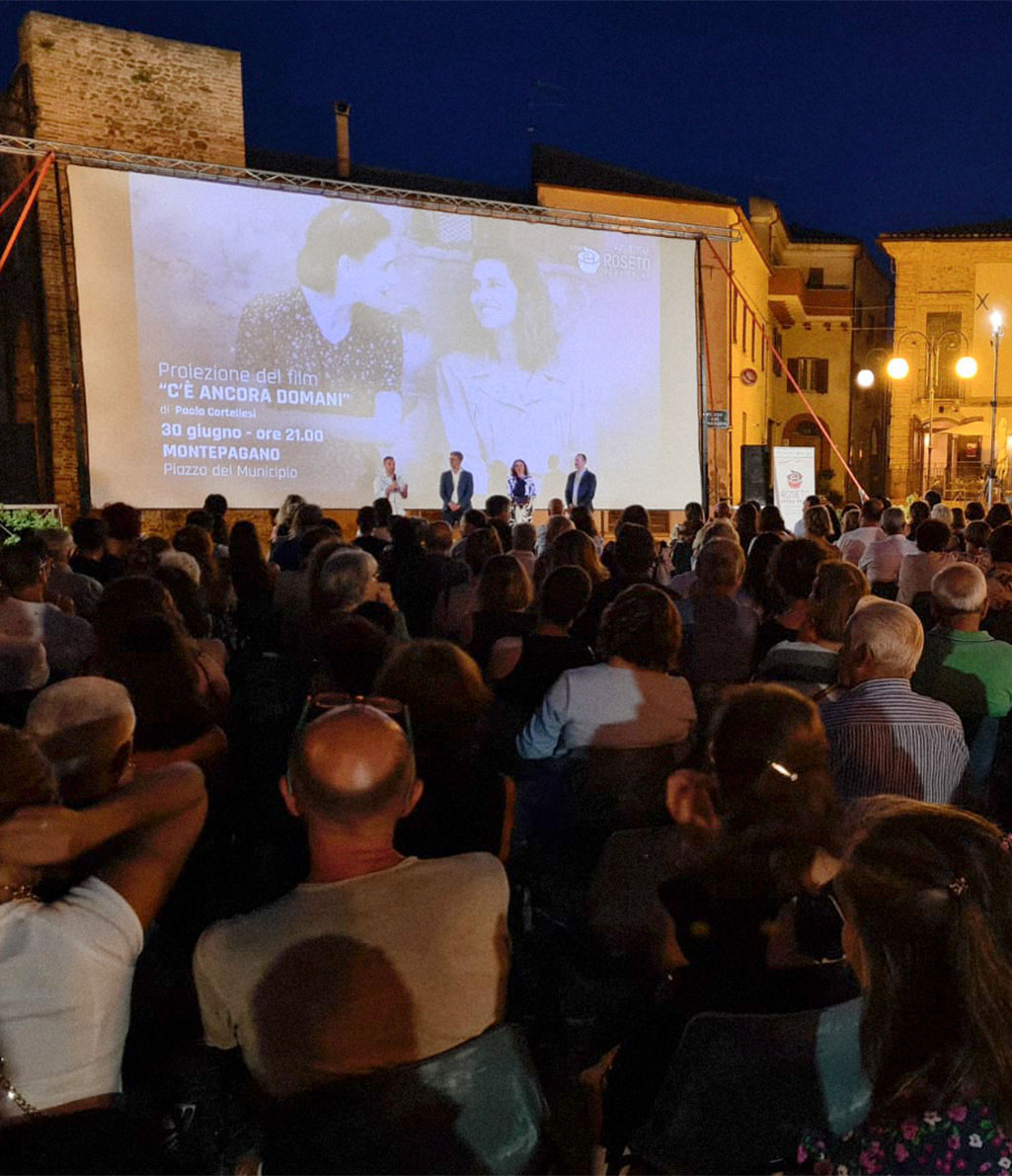 Un pubblico numeroso è seduto all'aperto di notte, guardando un grande schermo su cui viene proiettato il film "C'È ANCORA DOMANI". Sul palco davanti allo schermo, tre persone sono sedute a un tavolo, mentre altre due sono in piedi. L'evento si svolge in una piazza cittadina con edifici storici illuminati sullo sfondo.