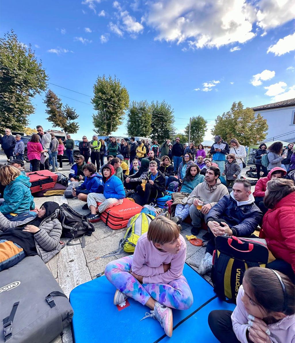 Un ampio gruppo di persone è seduto e in piedi all'aperto, molti con zaini e attrezzatura da arrampicata come crash pad, sotto un cielo blu con nuvole sparse. Sembrano partecipare a un evento o una gara sportiva.