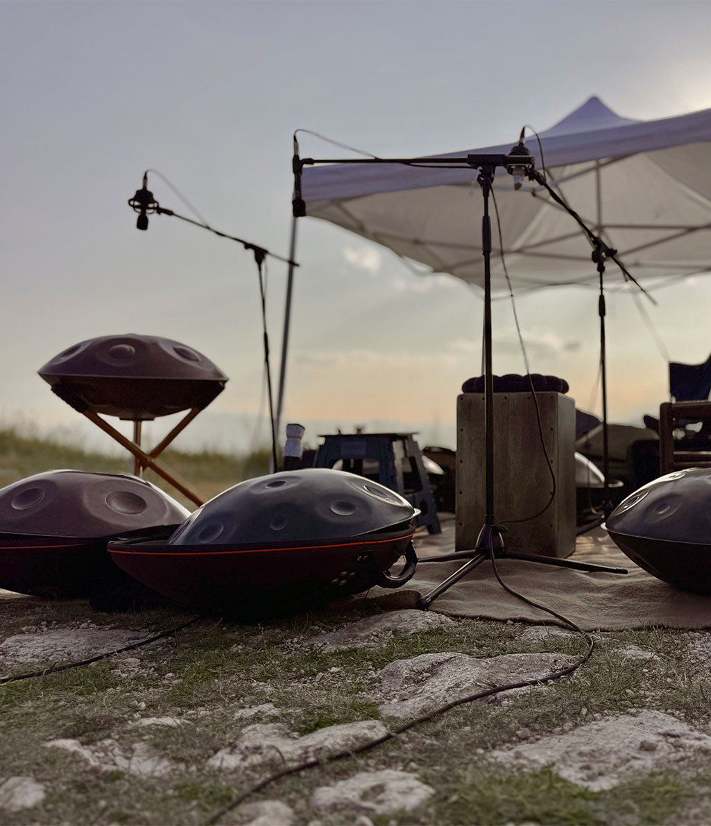 Un allestimento musicale all'aperto, probabilmente su una spiaggia o in un'area sabbiosa. Diverse handpan sono posizionate sul terreno e su un supporto in legno, con microfoni e stativi. Dietro, si intravede una tenda bianca e il cielo al tramonto.