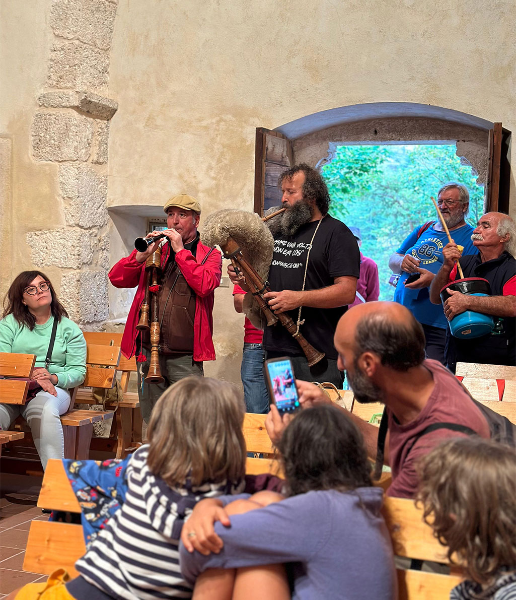 Un gruppo di musicisti folk sta suonando strumenti tradizionali, inclusa una zampogna, all'interno di un edificio con muri in pietra e aperture ad arco. Diverse persone assistono all'esibizione, alcune sedute su panche di legno, altre in piedi o chinate a guardare, con un uomo in primo piano che scatta una foto con il suo telefono.