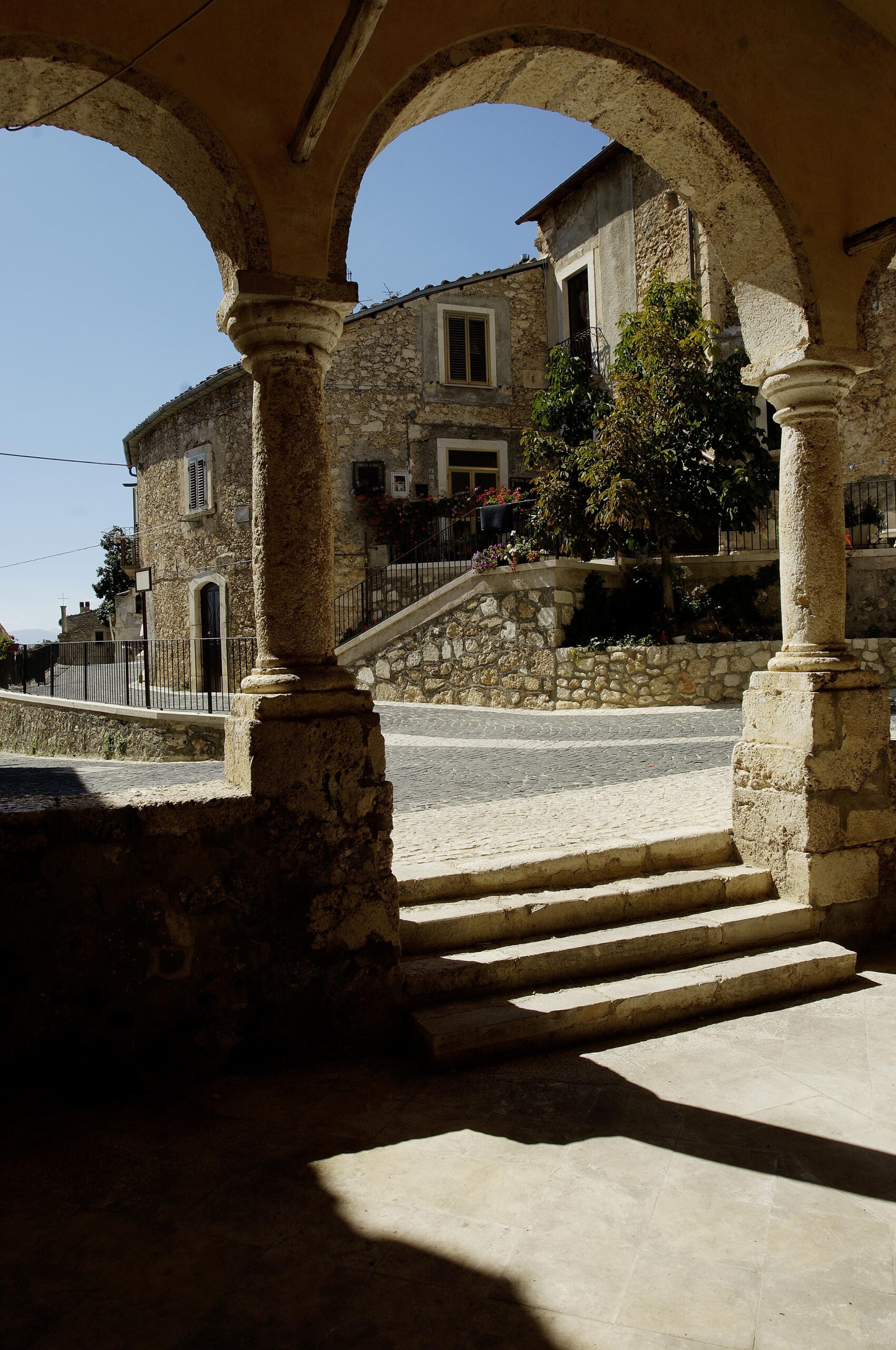 Una vista attraverso un porticato con archi e colonne in pietra che incorniciano una piazza acciottolata e un edificio tradizionale in pietra con scale e un albero verde, sotto un cielo limpido.