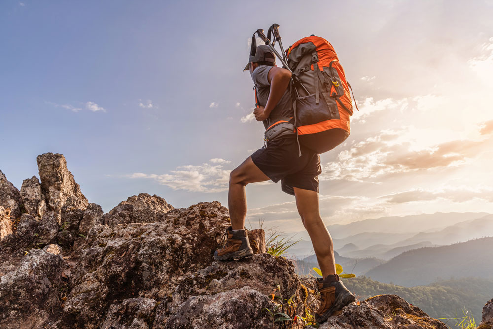 Escursionista con zaino grande e bastoncini da trekking che scala una roccia al tramonto, con vista panoramica sulle montagne.