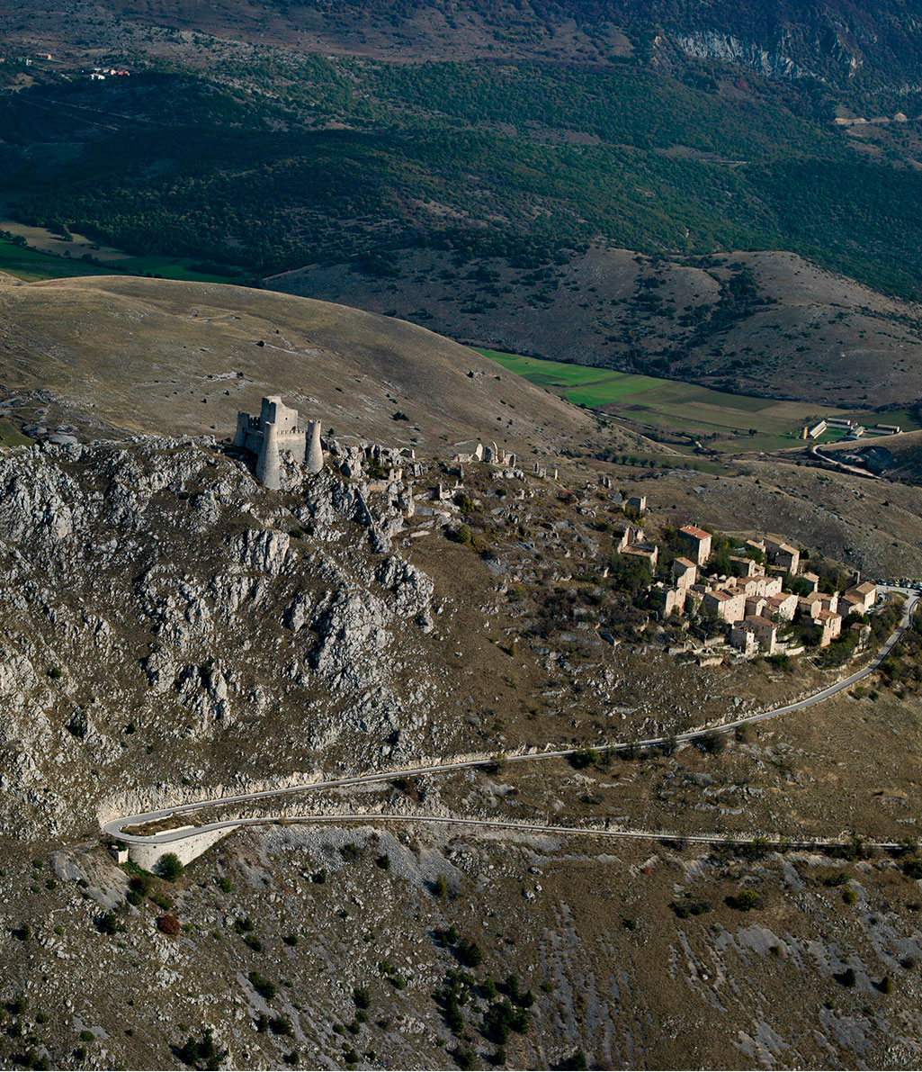Veduta aerea di un borgo medievale con un castello in cima a una collina rocciosa, collegato da una strada tortuosa che si snoda attraverso un paesaggio montano arido con valli verdi in lontananza.
