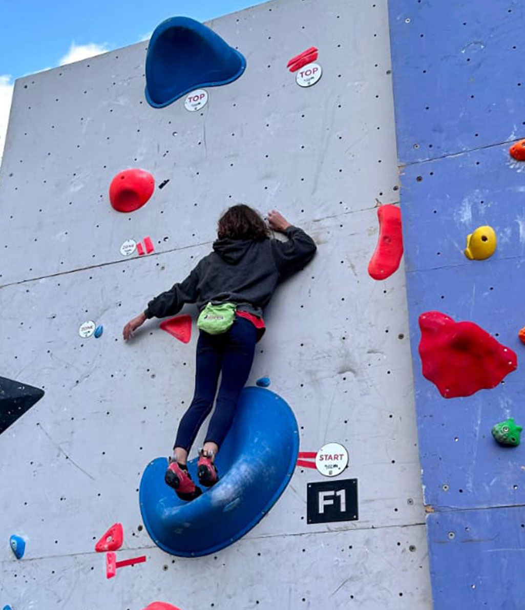 Una persona è intenta ad arrampicare su una parete artificiale da boulder di colore grigio, con prese rosse e blu. Un'altra parete di arrampicata blu con prese di vari colori è visibile sulla destra.