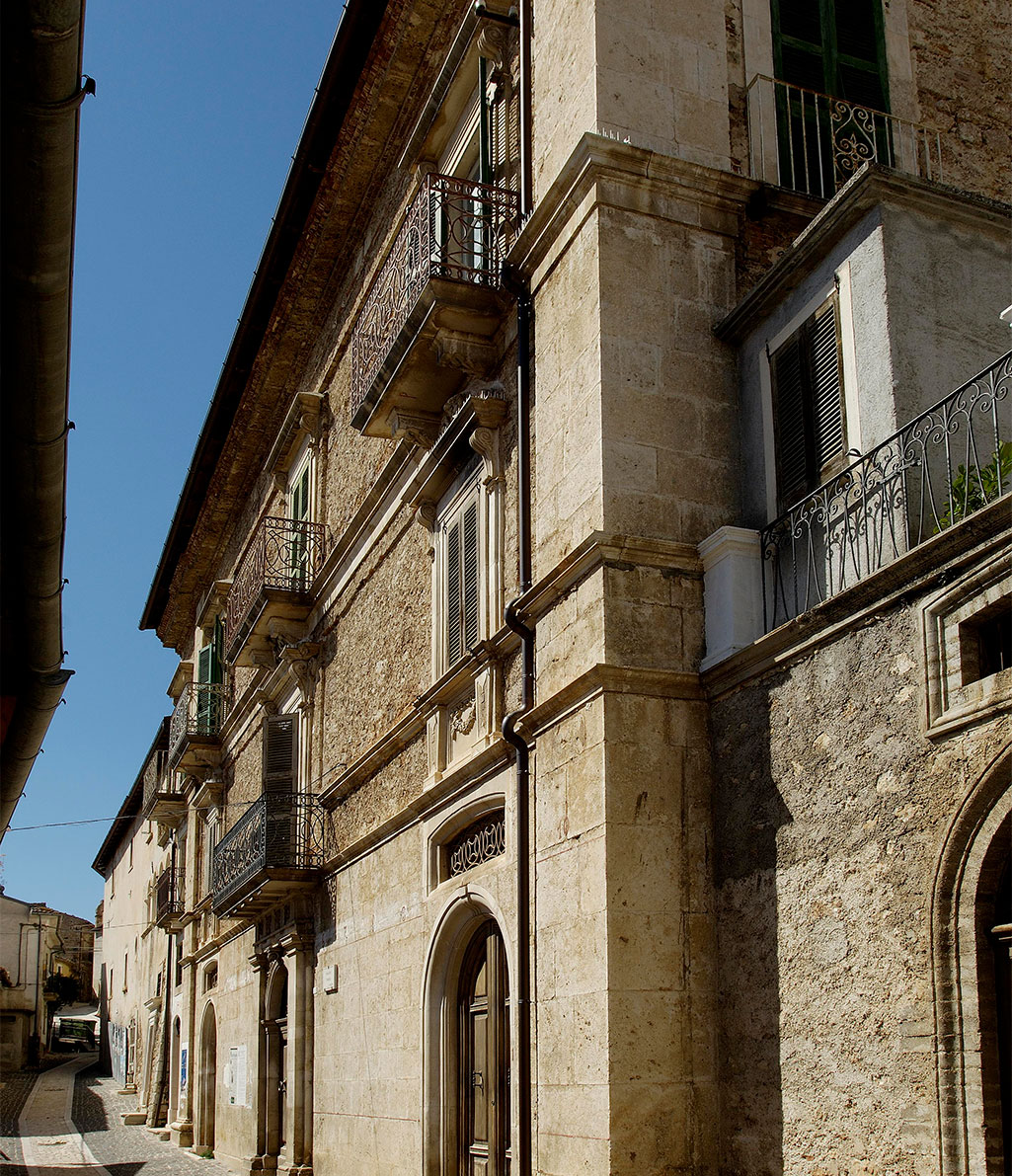 Una stretta strada fiancheggiata da imponenti edifici storici in pietra, con balconi in ferro battuto, persiane in legno e grandi portoni ad arco. Il cielo è limpido e blu.