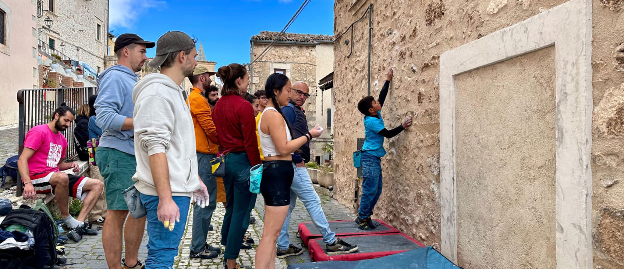 Un gruppo di persone osserva un bambino che scala una parete rocciosa urbana durante un evento di street boulder in un borgo antico.